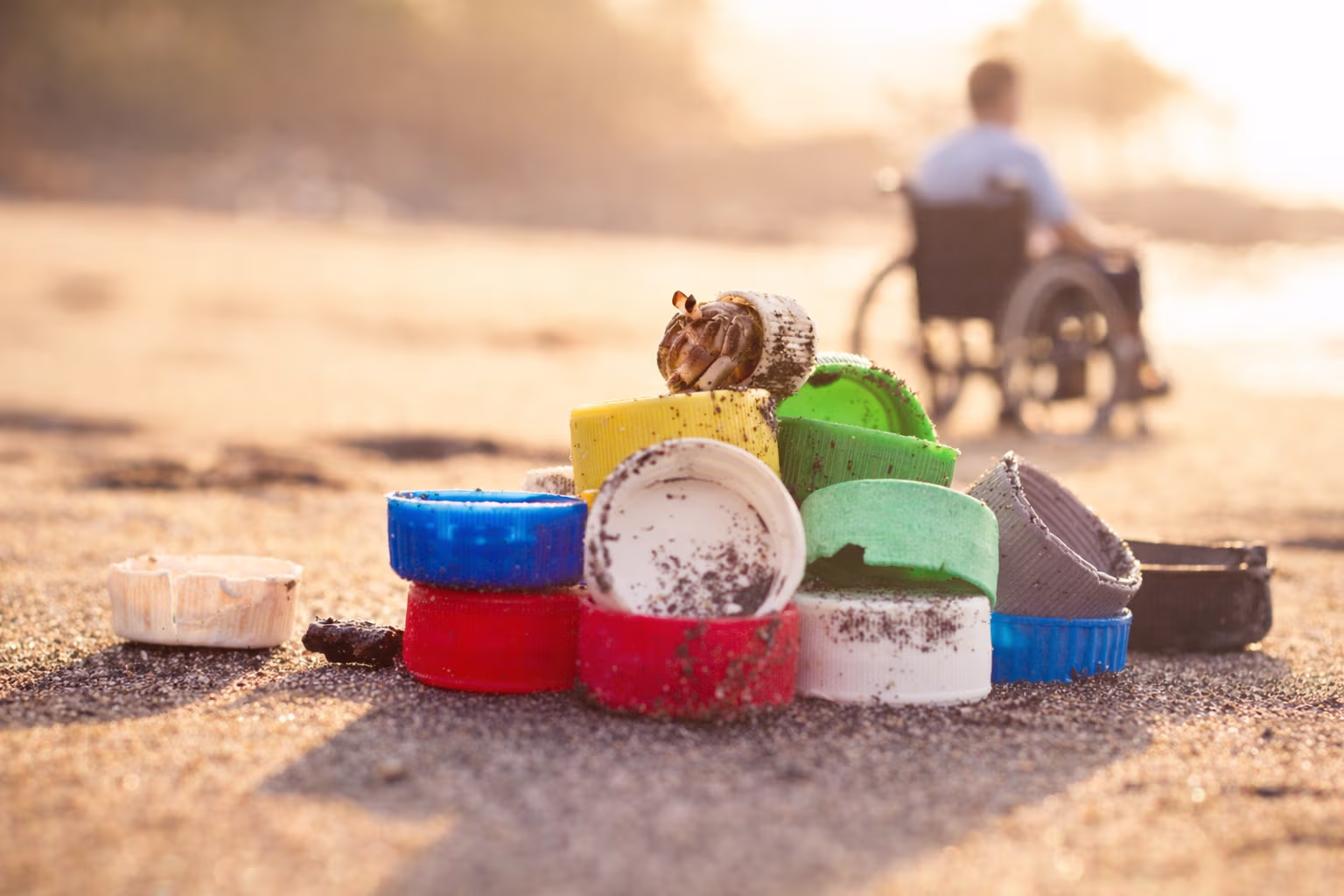 Plastikflaschendeckel mit einem Krebs daneben am Strand, im Hintergrund eine Person im Rollstuhl.