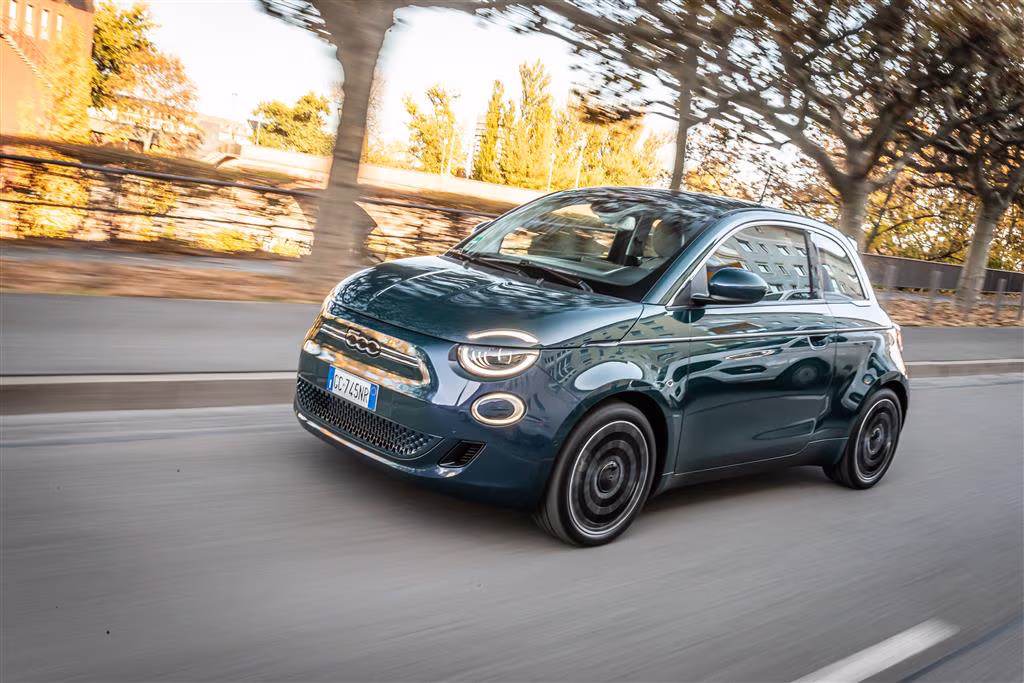 Blauer Fiat 500 Elektro fährt auf einer städtischen Straße mit Bäumen und Gebäuden im Hintergrund.