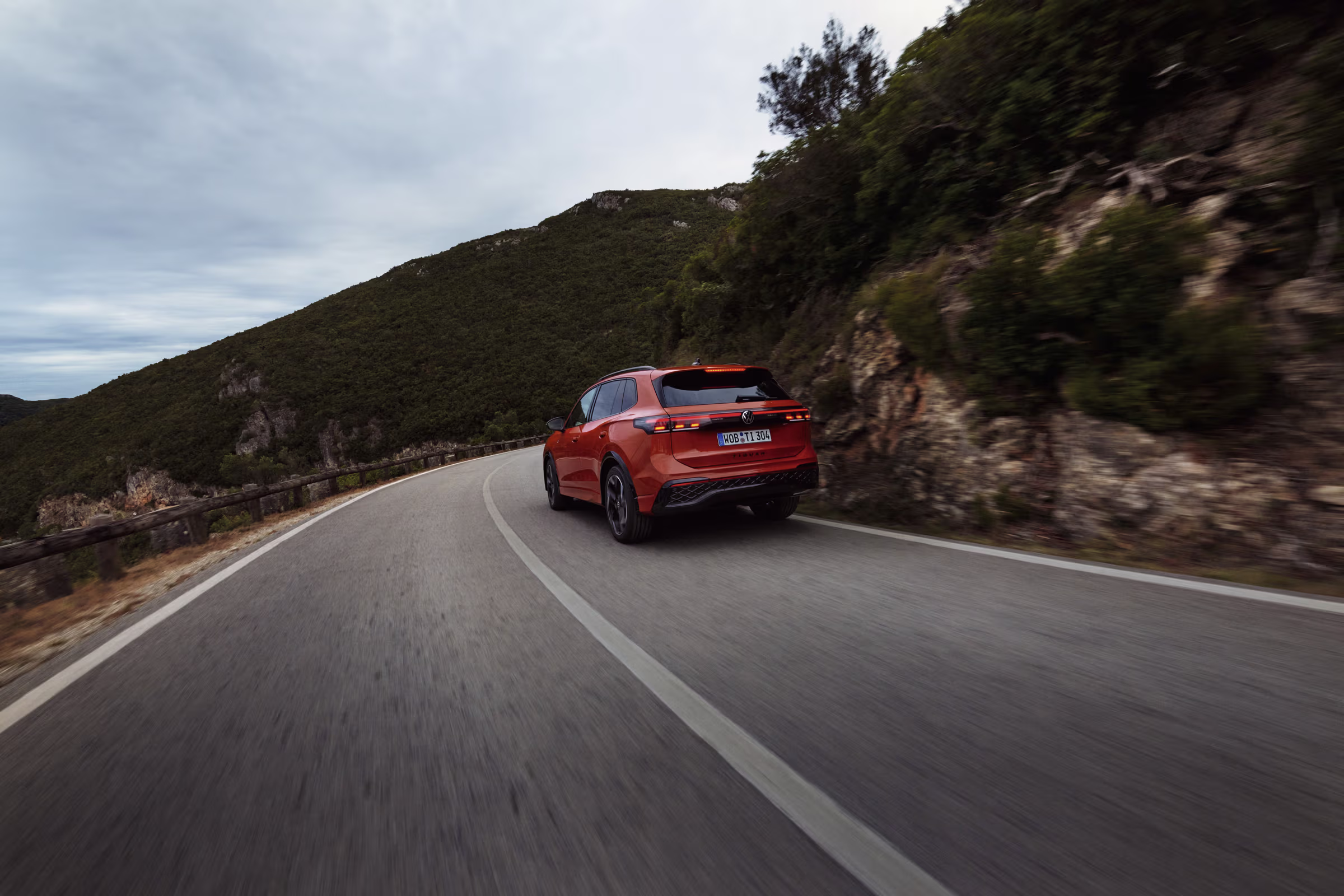 Rotes Auto fährt eine kurvenreiche Bergstraße entlang bei bewölktem Himmel.