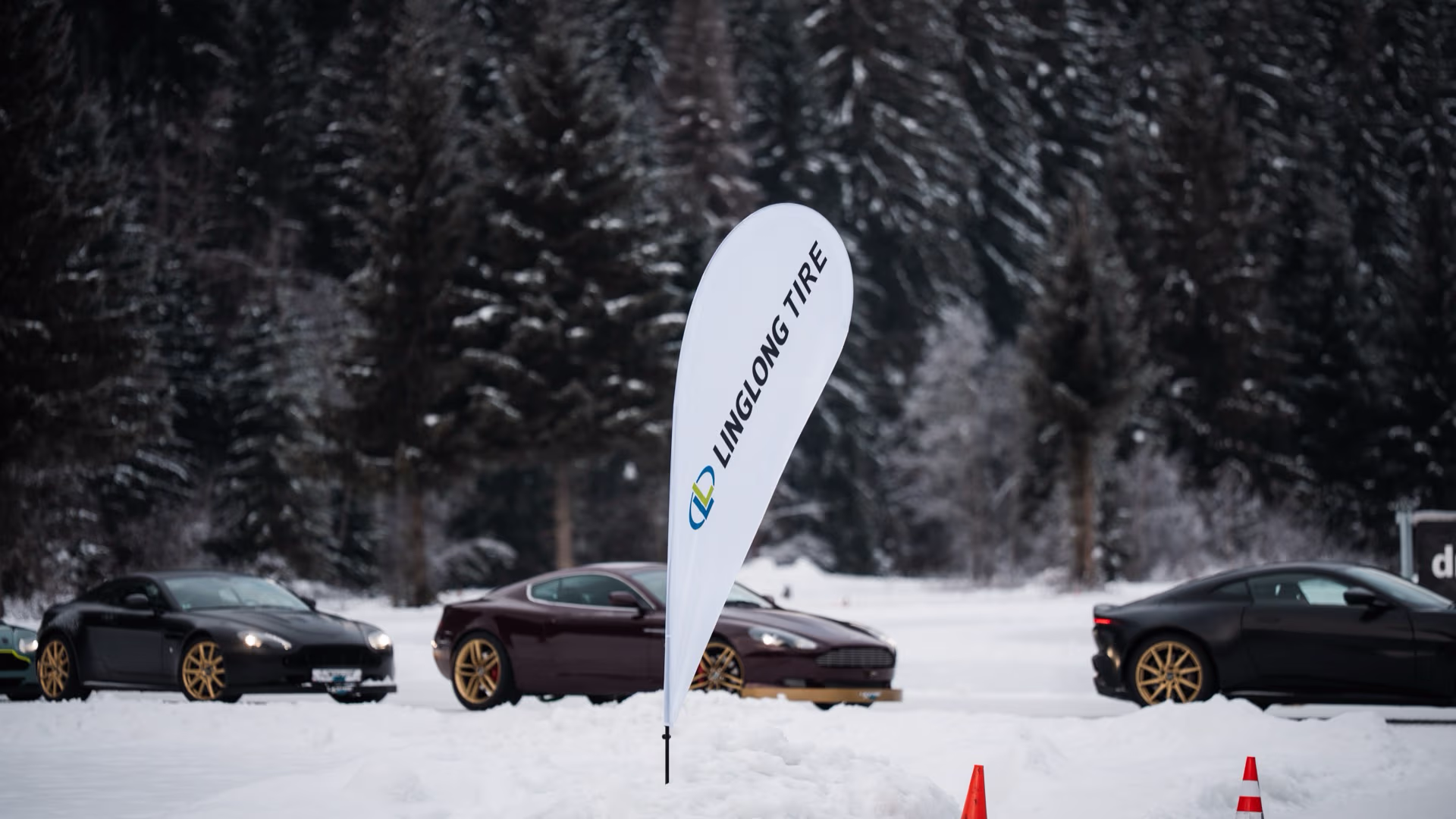 Drei sportliche Autos mit goldfarbenen Felgen parken auf schneebedecktem Gelände vor einem Wald mit Tannenbäumen; ein weißes, tropfenförmiges Banner mit dem Schriftzug ‚Linglong Tire‘ steht im Vordergrund.