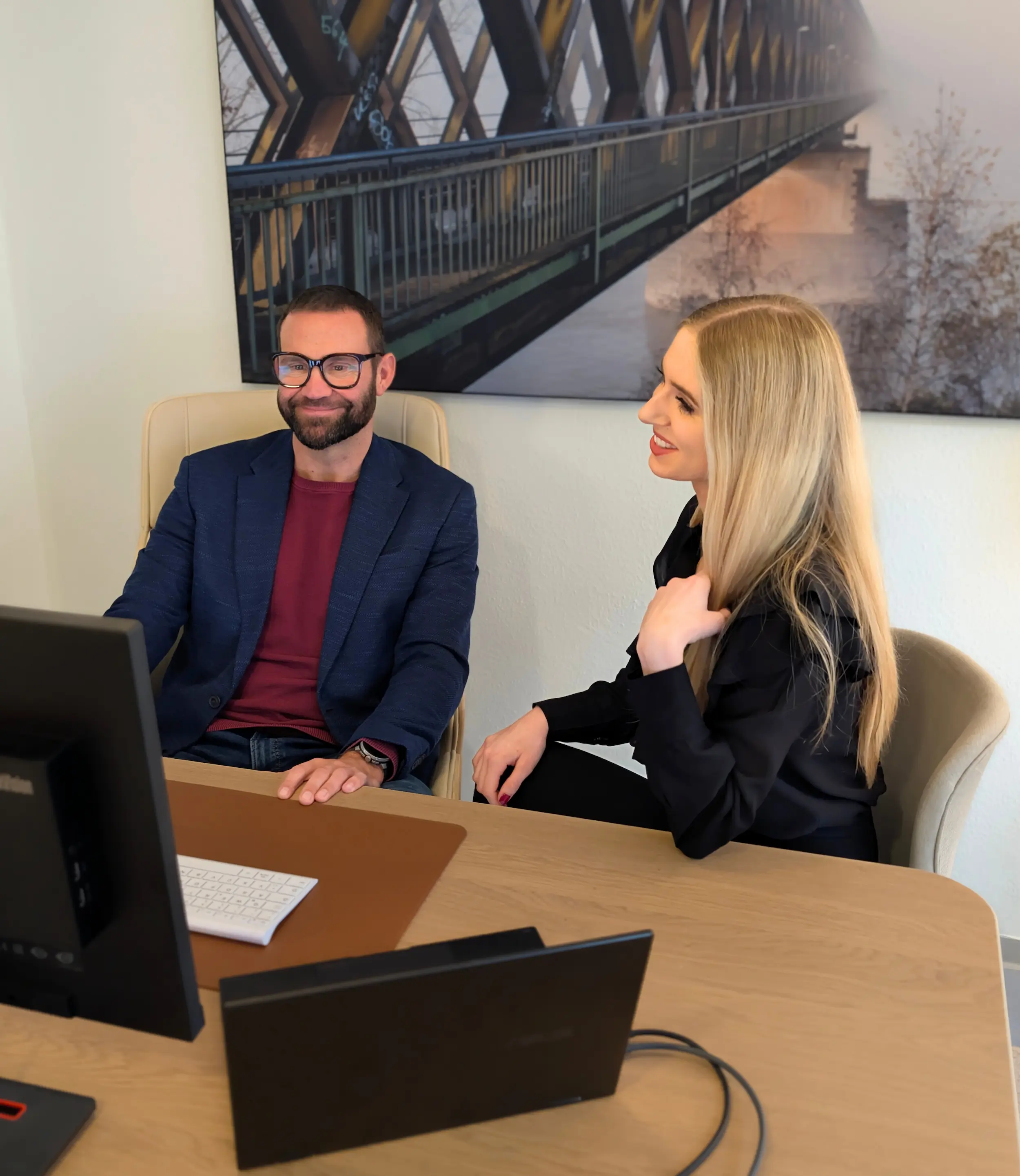 Ein Mann mit Brille und blauer Jacke sitzt an einem Schreibtisch neben einer lächelnden Frau mit langen blonden Haaren.