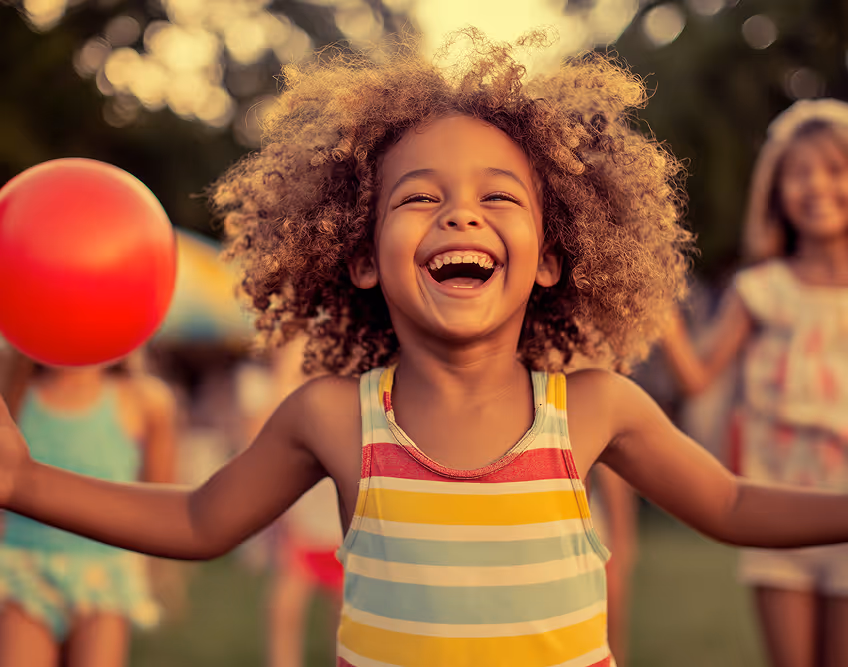 Smiling child with curly hair wearing a colorful striped tank top holding a red ball outdoors.