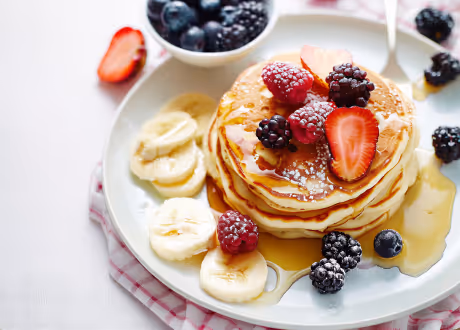 Stack of pancakes topped with syrup and mixed berries, including strawberries, raspberries, and blackberries, served with banana slices on a white plate.