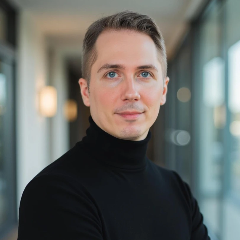 Portrait of a man with light skin, short light brown hair, and blue eyes wearing a black turtleneck sweater in a modern office hallway.