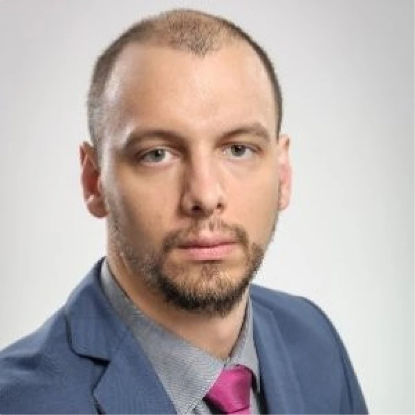 Serious man with short hair, beard, and mustache wearing a blue suit, gray shirt, and magenta tie against a light gray background.