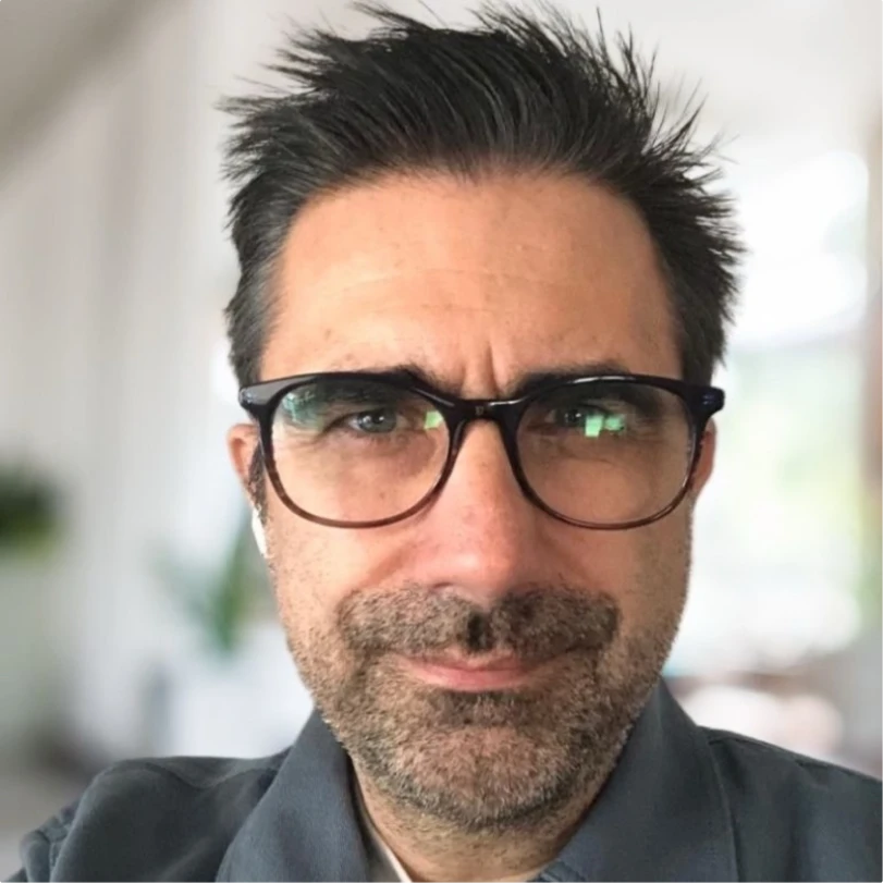 Close-up of a man with short spiky hair, glasses, and a slight beard, wearing a gray shirt, with a blurred indoor background.