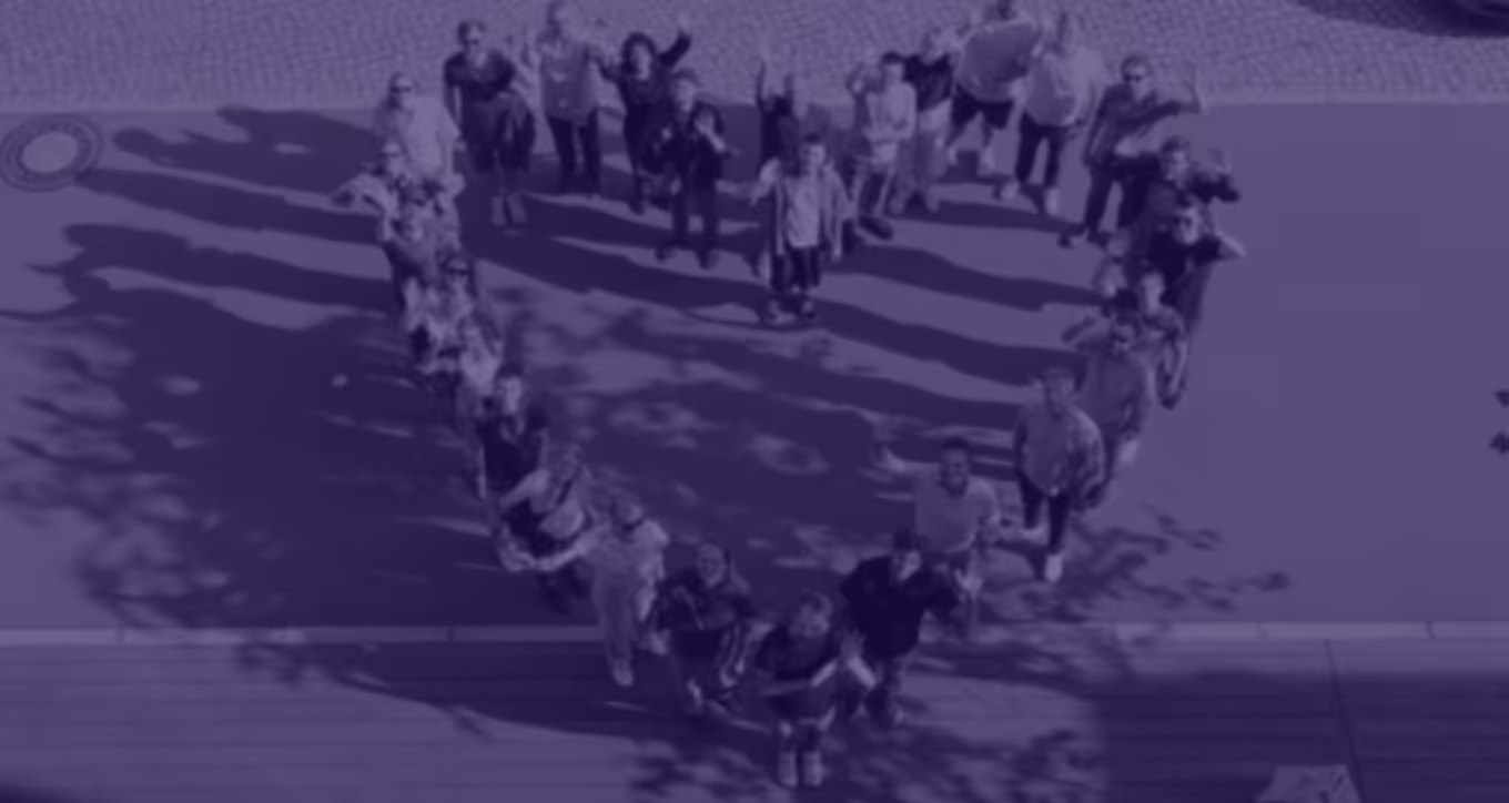 Eine Gruppe von Menschen steht im Freien in Form eines großen Herzens, schaut nach oben und winkt.