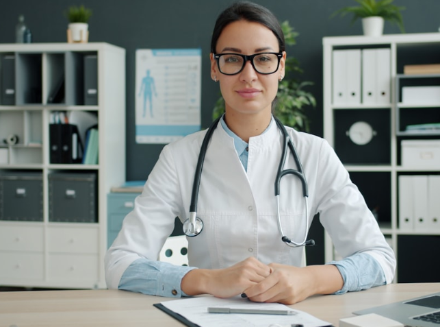 Female doctor wearing glasses and a white coat with a stethoscope around her neck sitting at a desk with medical documents.