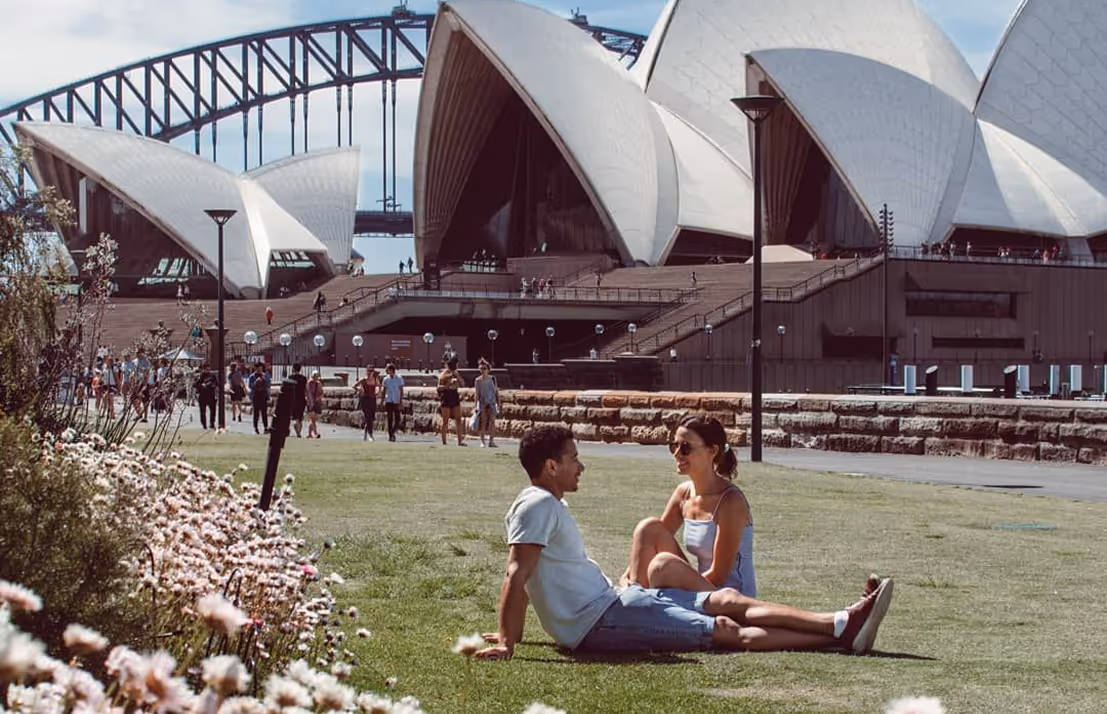 Couple sitting on grass near Sydney Opera House with Sydney Harbour Bridge in the background.