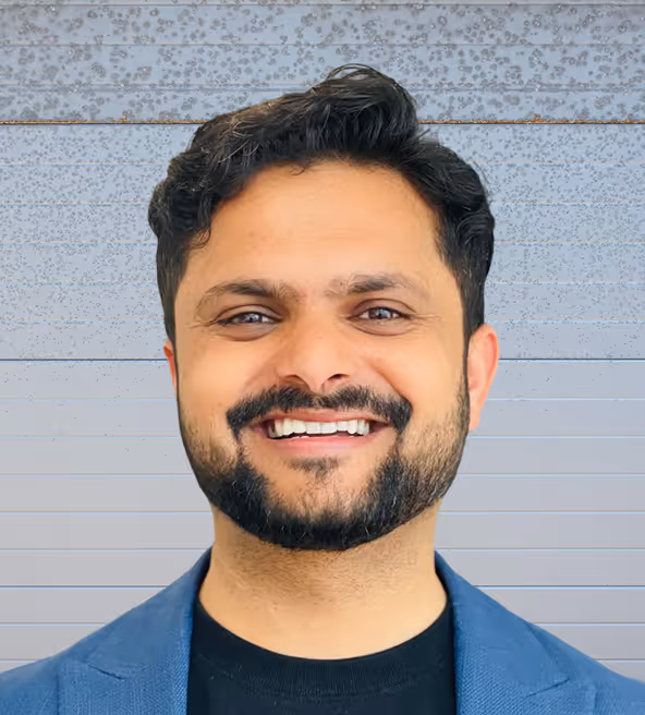 Smiling man with black hair and beard wearing a blue blazer and black shirt against a grey textured background.