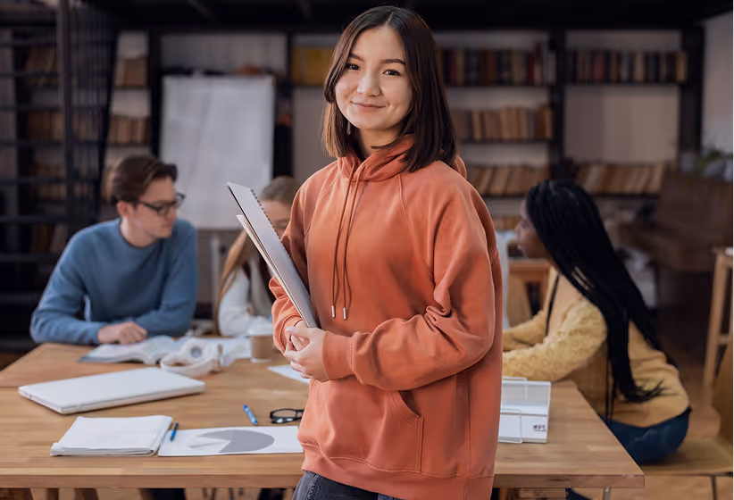 Young woman in an orange hoodie holding a folder, smiling at the camera with three people studying at a table in the background.