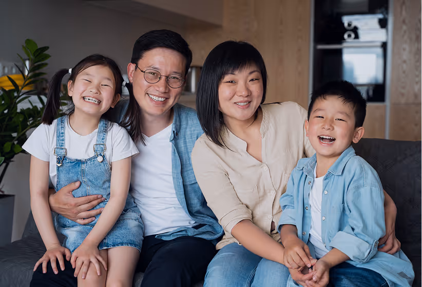 Smiling Asian family of four sitting closely on a couch in a warmly lit living room.