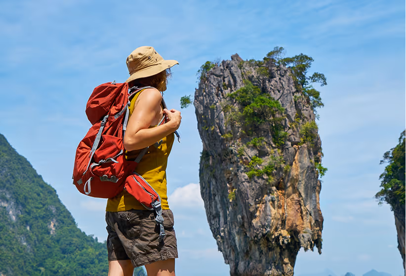 Woman with a red backpack and hat hiking near a tall limestone rock formation with greenery under a blue sky.