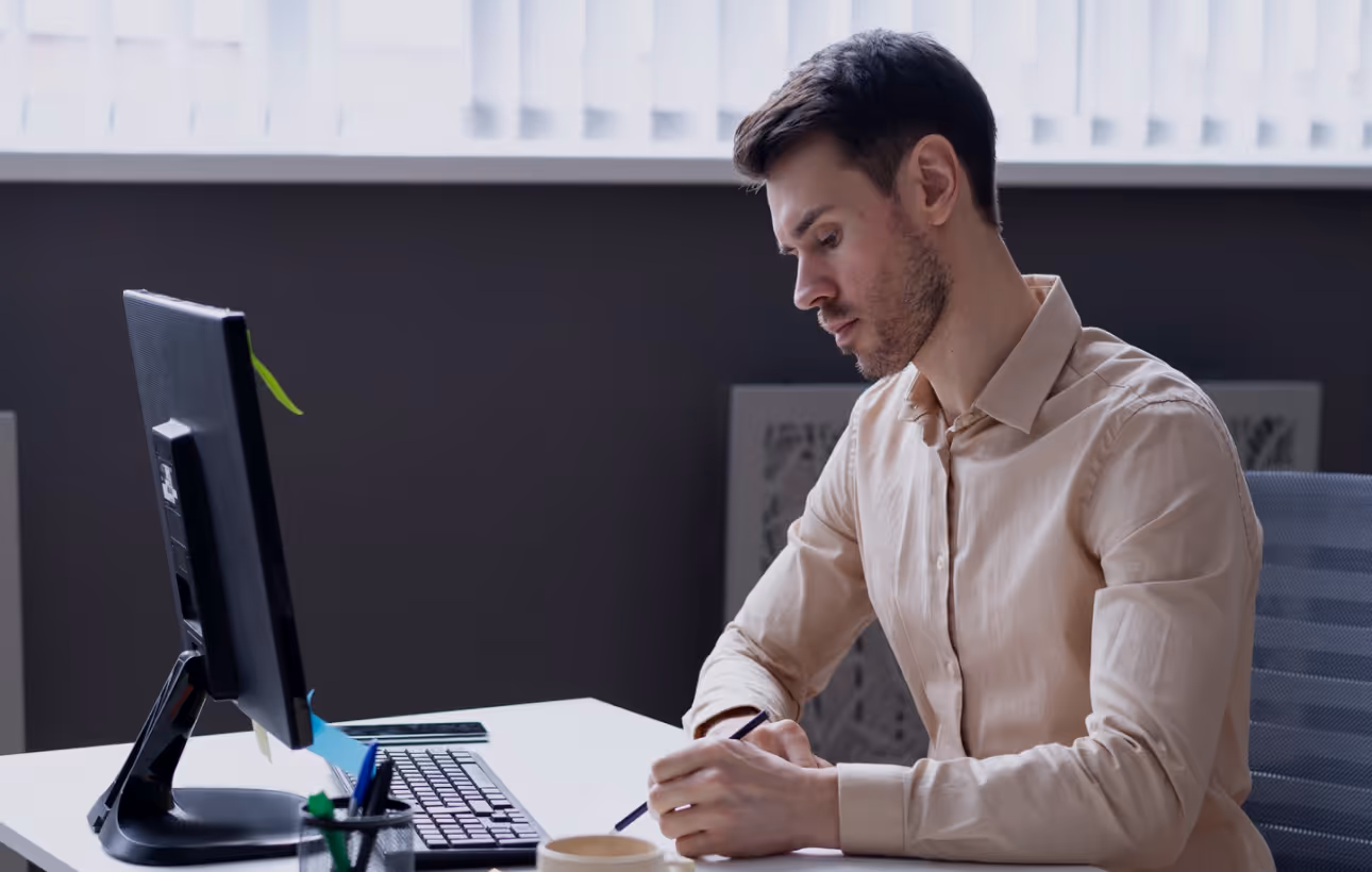 Man in beige shirt sitting at desk and writing with pen in office setting.