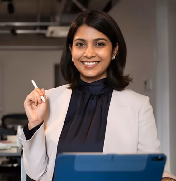 Smiling woman with shoulder-length black hair in a light blazer and black blouse holding a stylus, sitting behind a tablet.
