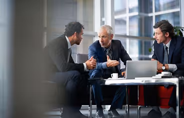 Three men in business attire engaged in a discussion around a table in a modern office.