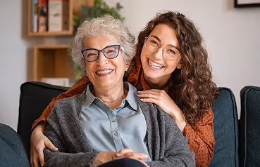 Smiling elderly woman with gray hair and glasses sitting on a couch with a younger woman hugging her from behind.