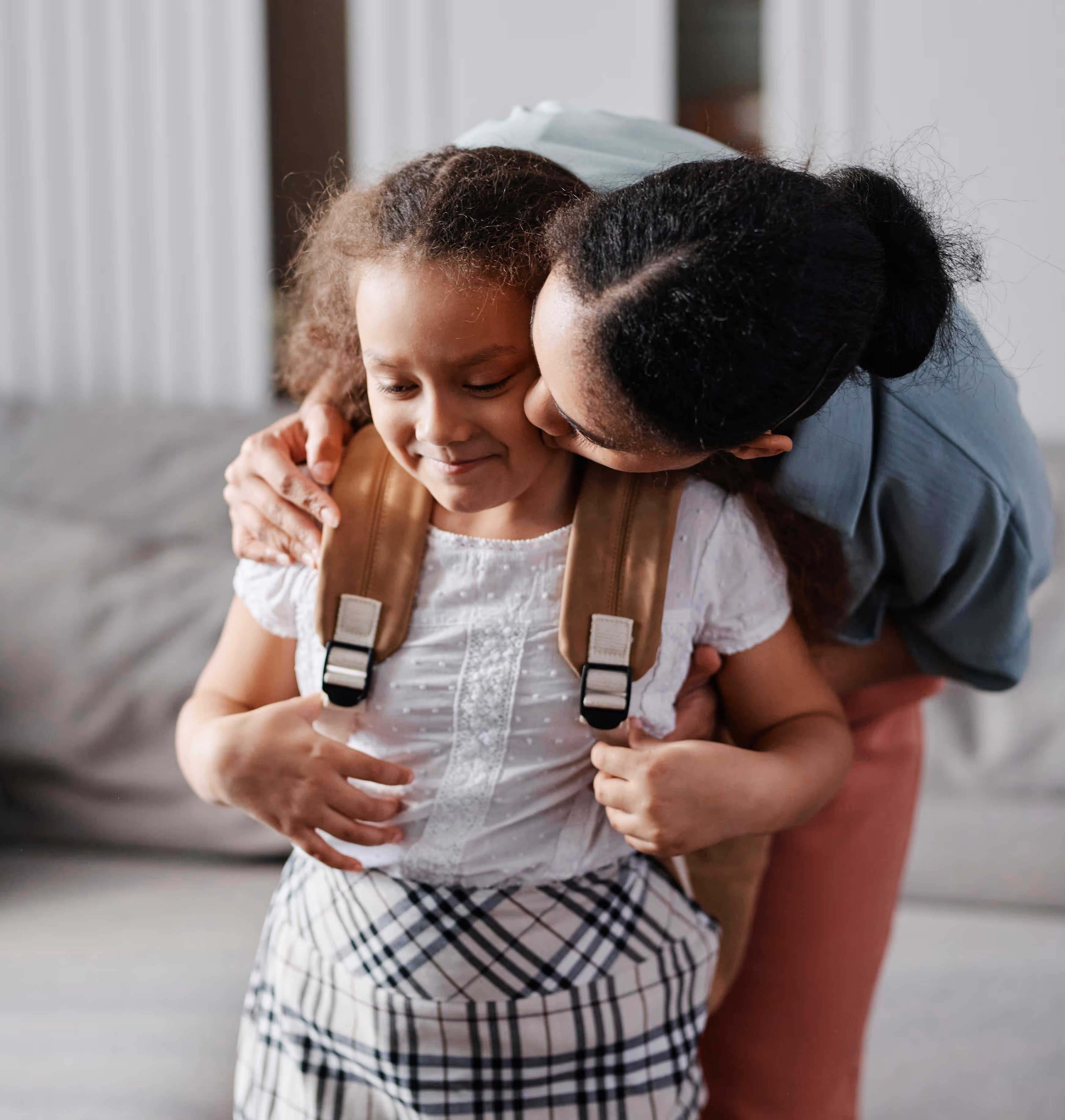 Dependent child standing indoors with a backpack while being embraced by an adult.