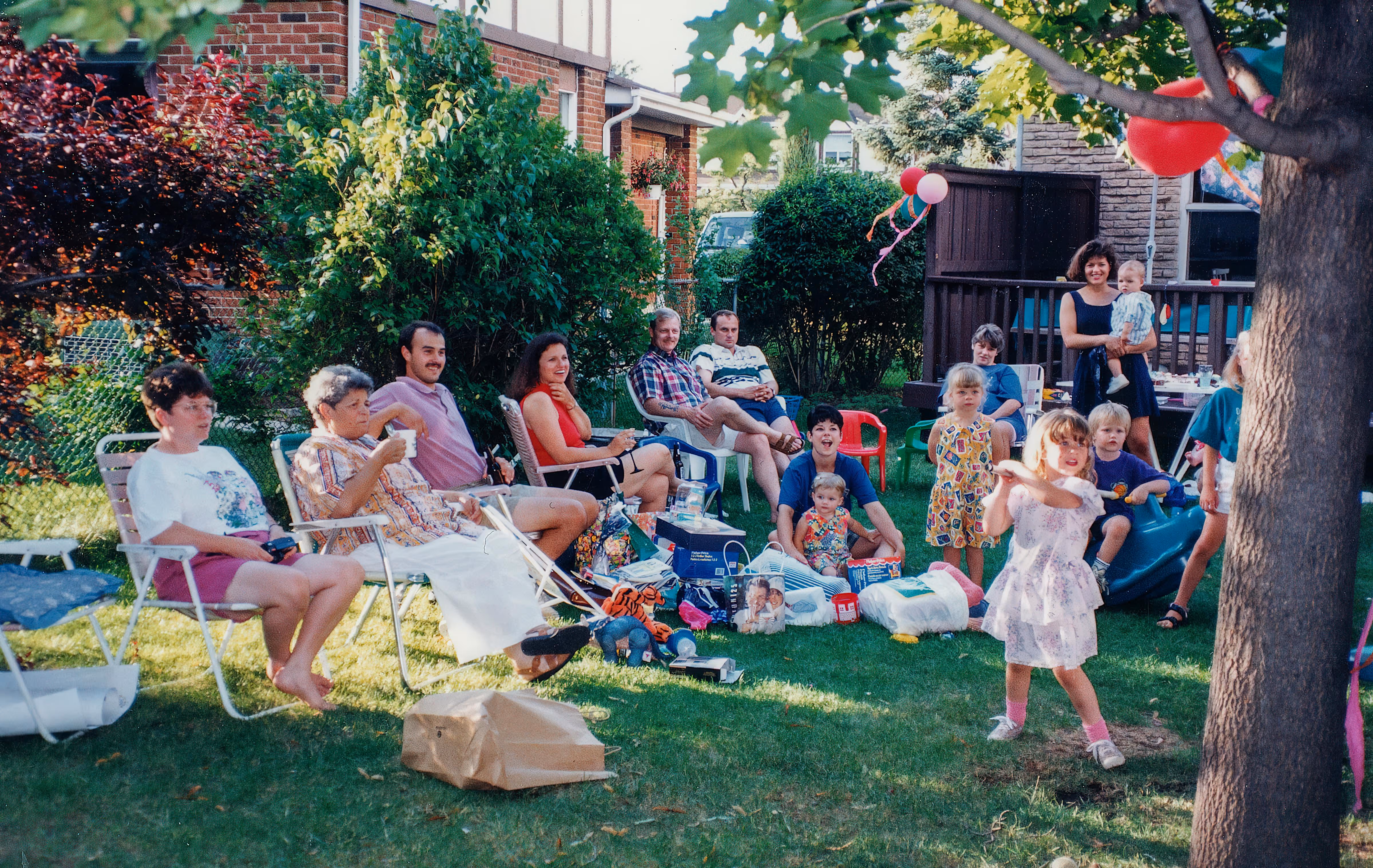 Relatives gathered outdoors in a backyard with adults seated on lawn chairs and children playing nearby.