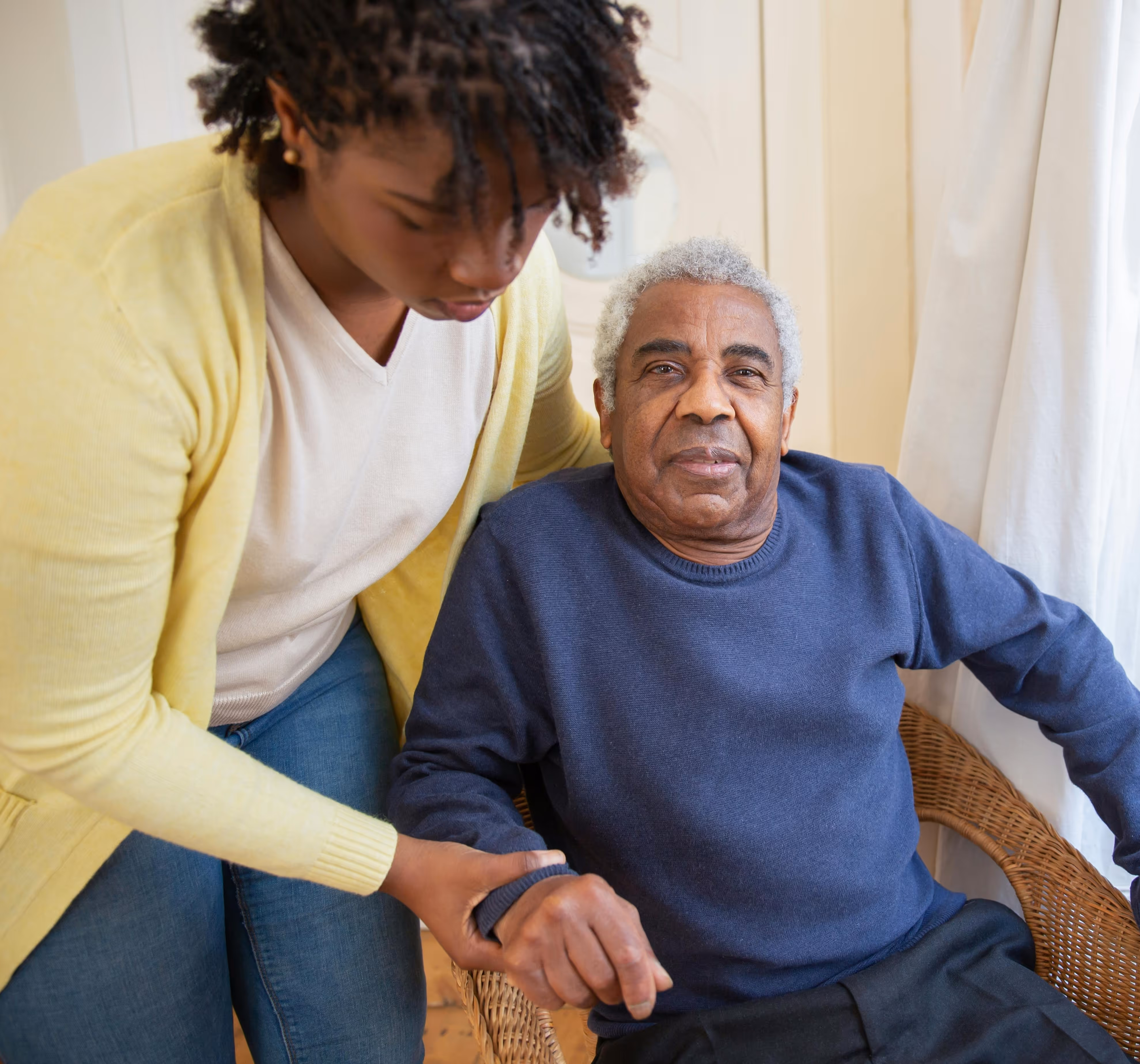 Carer assisting an older adult indoors for a carer visa application while supporting him from a chair.