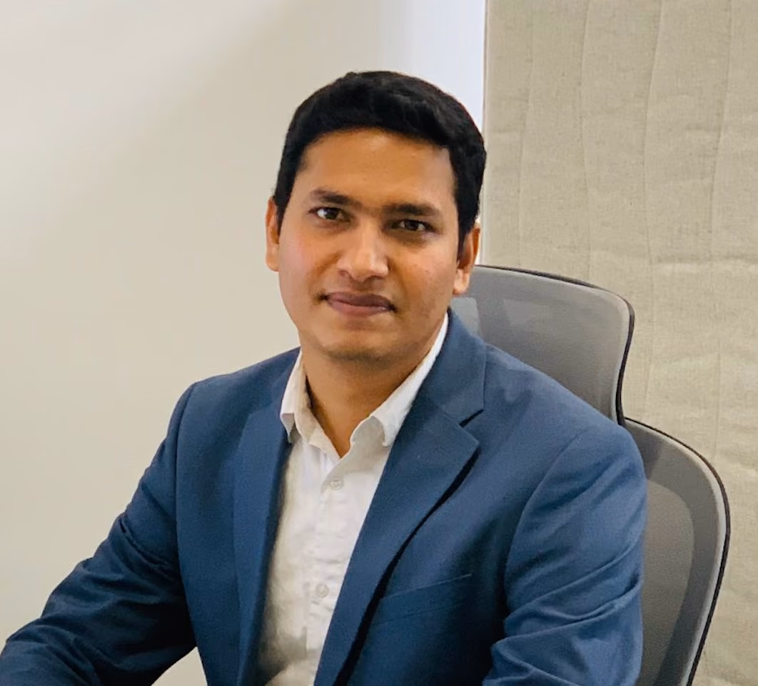 Man in a blue blazer and white shirt sitting in an office chair against a plain wall.