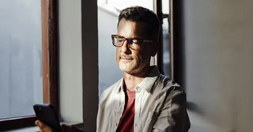 Man wearing glasses and a light shirt looking at a smartphone indoors near a window with sunlight on his face.