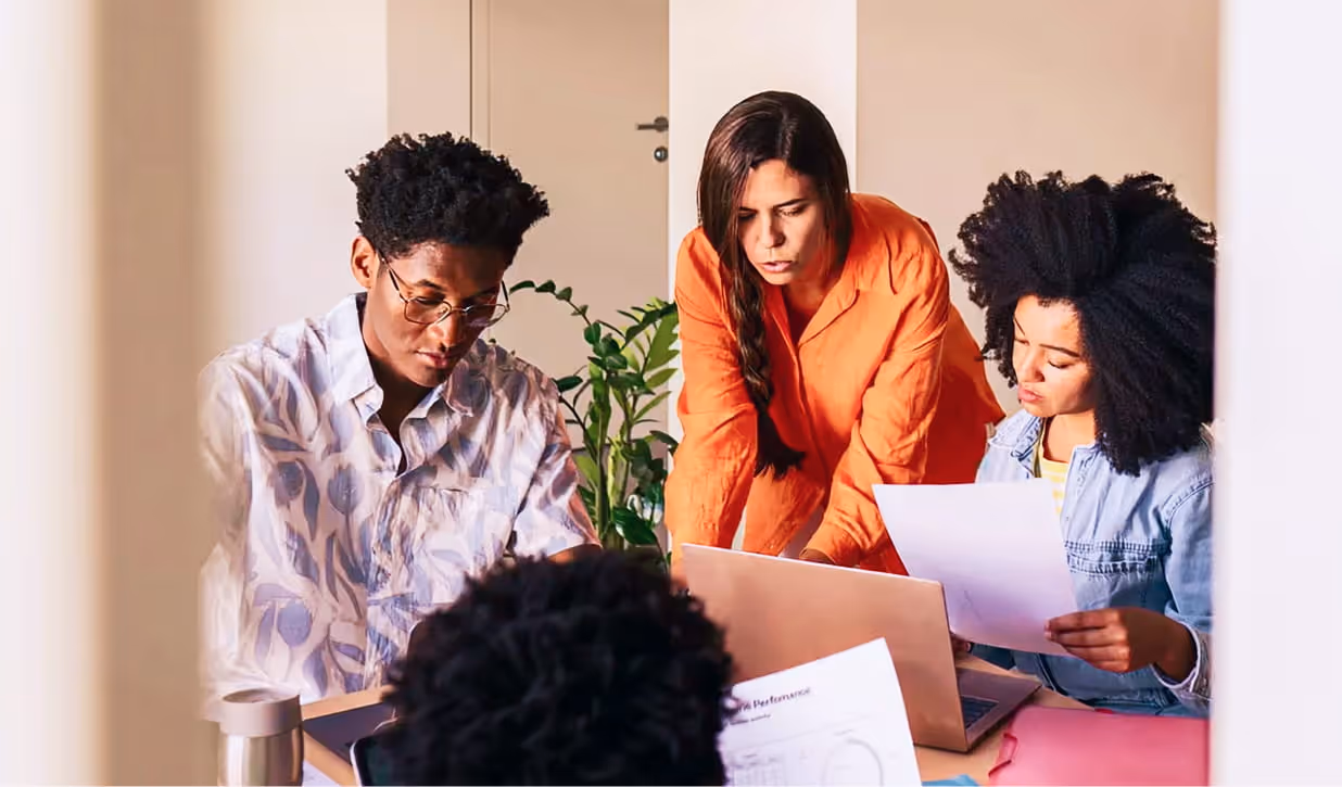 Three young adults collaborating around a laptop and documents in a bright meeting room.
