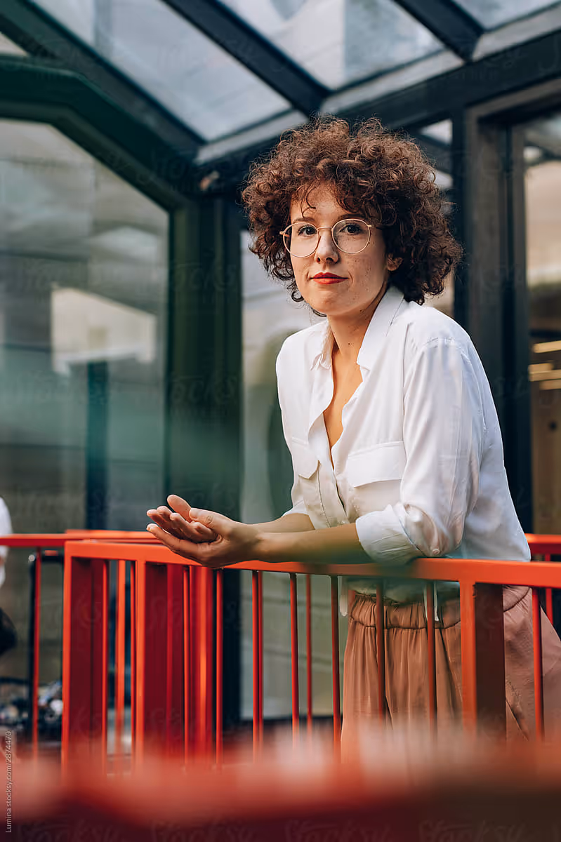 Smiling young professional woman with glasses and curly hair, leaning on a red railing in a bright, modern office setting.