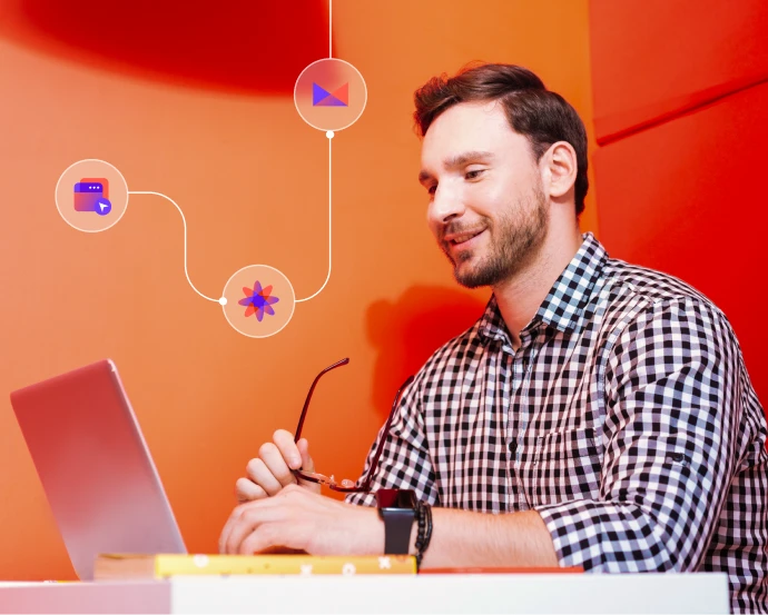 Man in checkered shirt sitting at a desk using a laptop with digital icons floating above him.