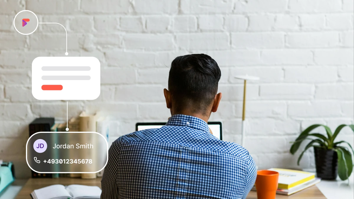 Man in blue checked shirt working on laptop at desk with books, plant, and orange cup nearby.