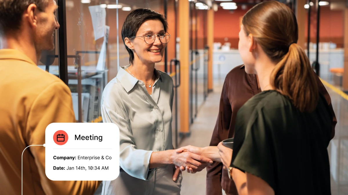 Two women shaking hands in an office hallway while two other colleagues stand nearby.