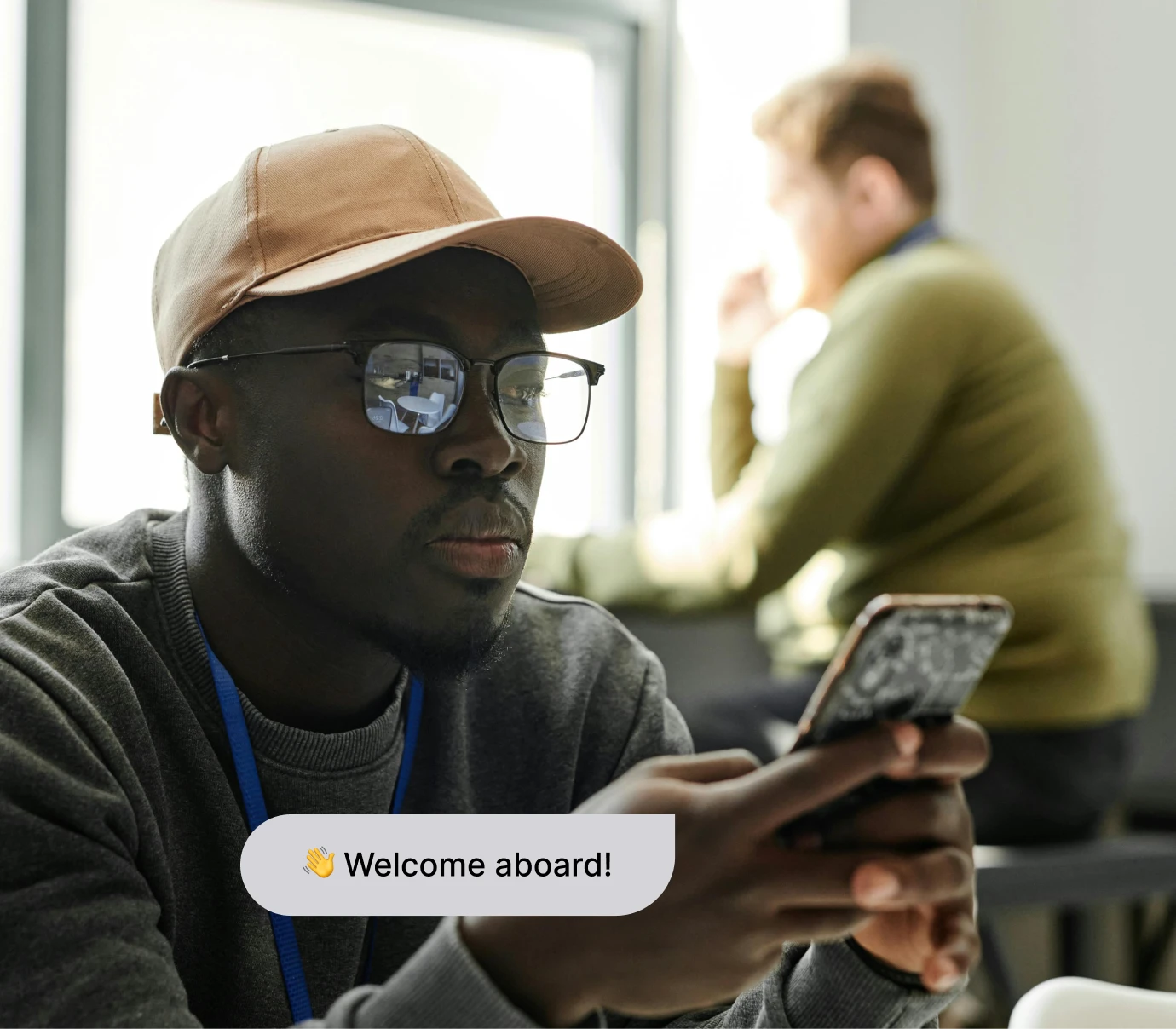 Man wearing glasses and cap looking at smartphone with another man blurred in background.
