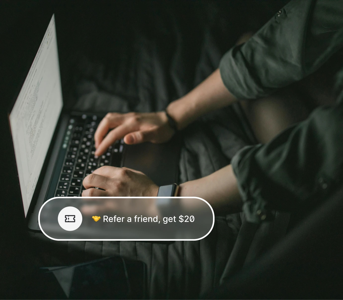 Person typing on a laptop in a dark room with a referral offer overlay saying 'Refer a friend, get $20'.