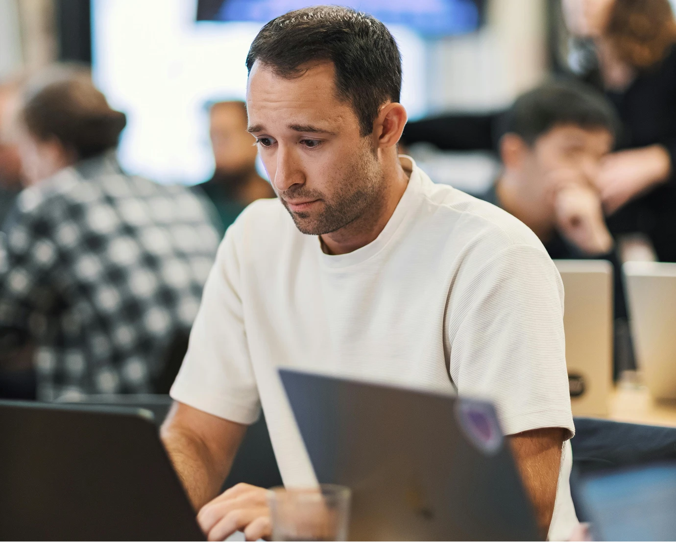 Man in white shirt focused on typing on laptop in a busy office with others working in the background.