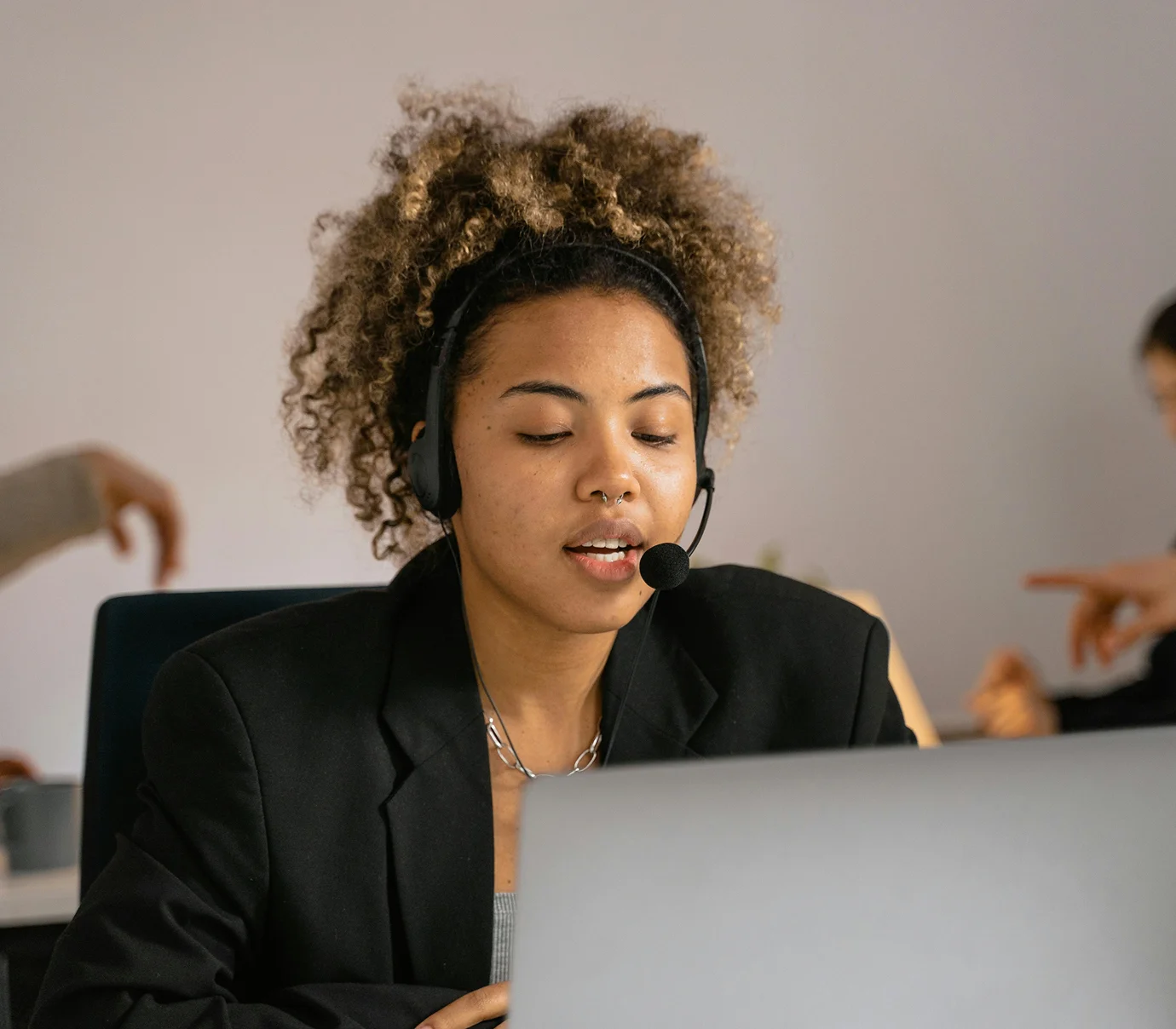 Young woman with curly hair wearing headset and speaking while looking at a laptop screen.