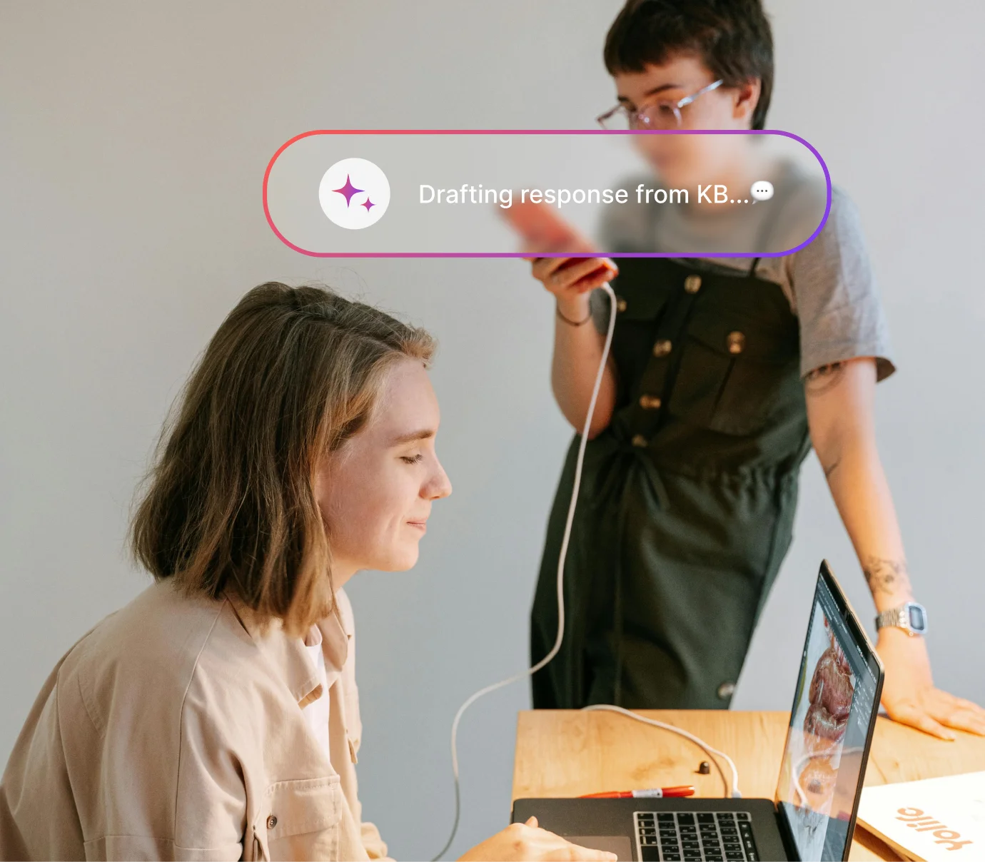 Two people working together, one on a laptop and another using a mobile phone with charging cable connected.