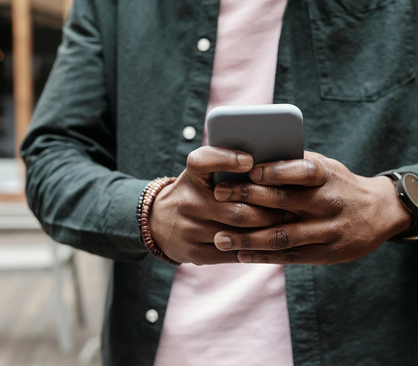 Close-up of a person holding and using a smartphone with both hands, wearing a dark shirt and bracelets.