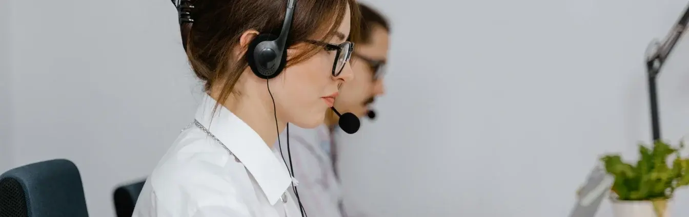 Two call center agents wearing headsets and glasses working at computers.