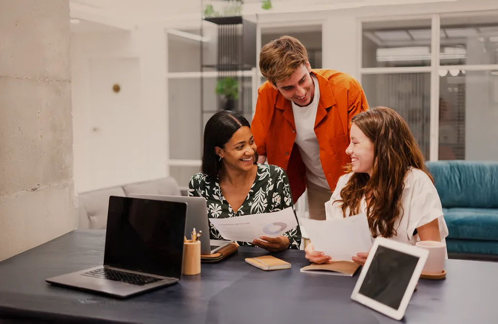 Three coworkers smiling and reviewing documents together at a desk with laptops and tablet.