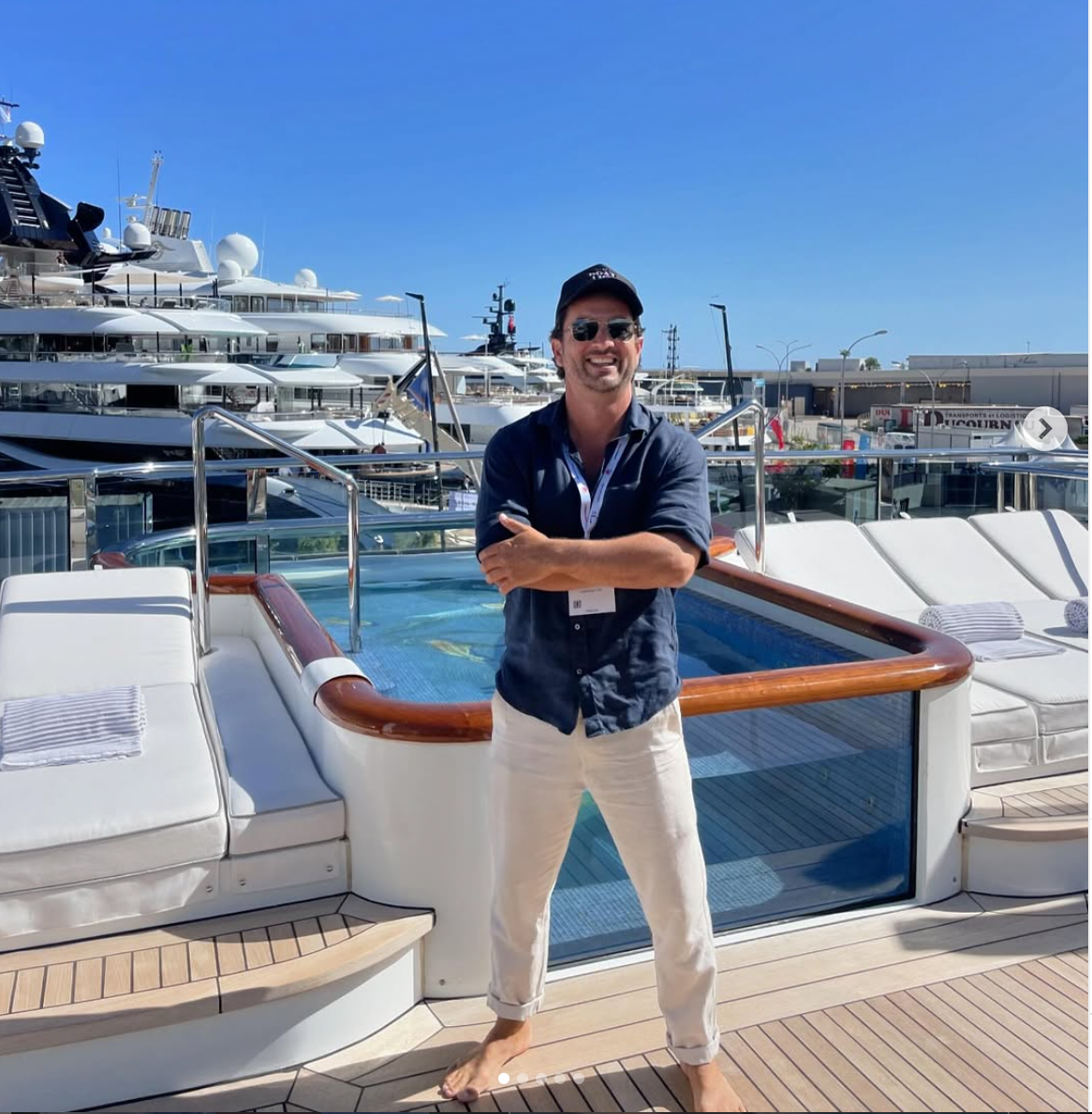 Man wearing sunglasses, a dark shirt, and white pants standing barefoot on a yacht deck with a pool and other yachts in the background.