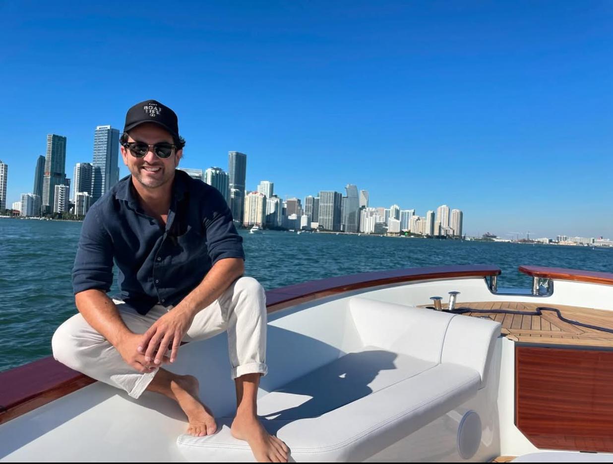 Man in sunglasses and cap sitting barefoot on the bow of a boat with a city skyline across the water under a clear blue sky.