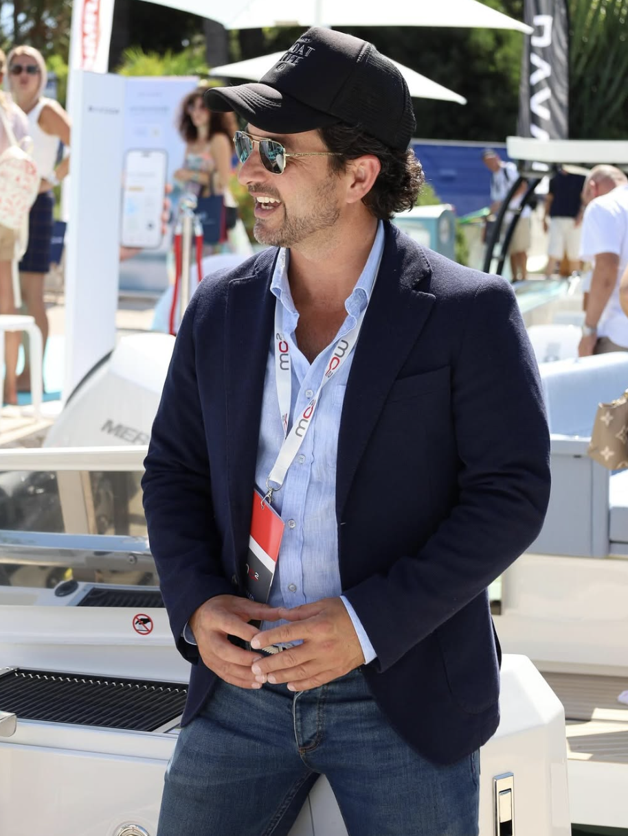 Man wearing a black cap, sunglasses, blue blazer, and jeans standing and smiling outdoors at a boat show.