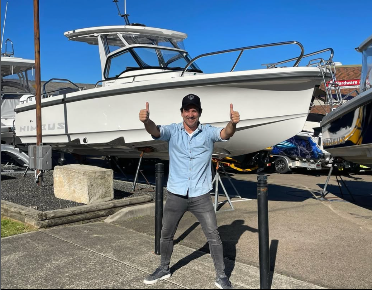 Man in casual clothes and cap standing on pavement with thumbs up in front of a white Nimbus motorboat on display.