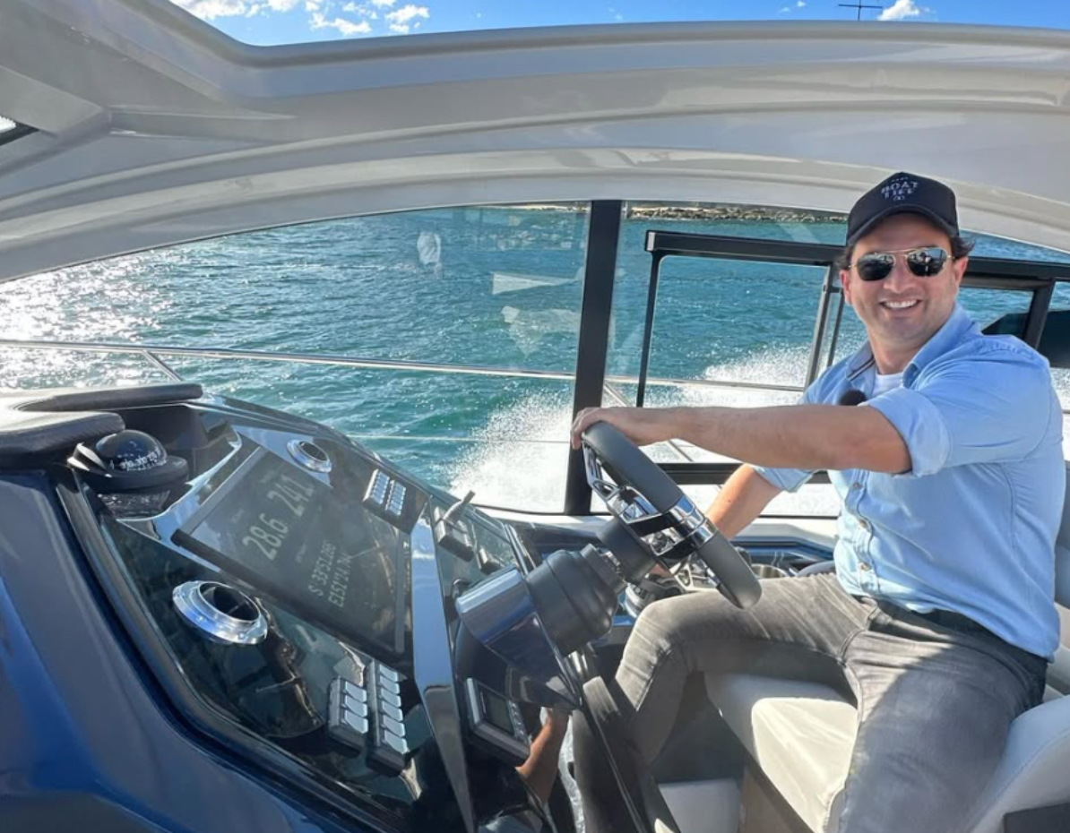 Man wearing sunglasses and a cap steering a boat on a sunny day with water visible through the windows.
