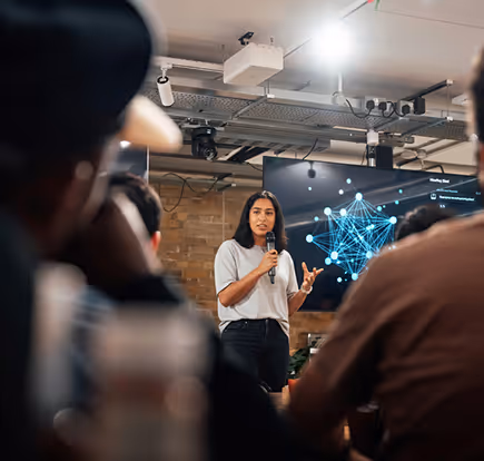 Woman holding a microphone presenting data visualization on a screen to an audience in a modern meeting room.