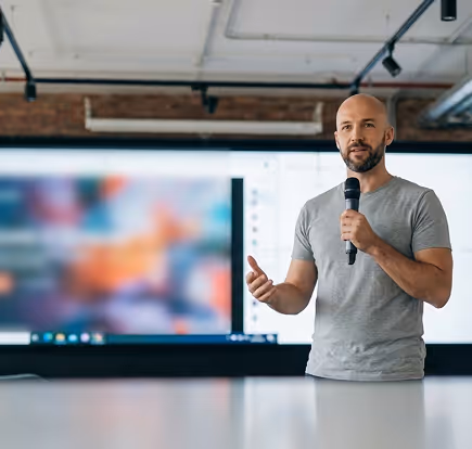 Man with a microphone giving a presentation in front of a large blurred screen in an industrial-style room.