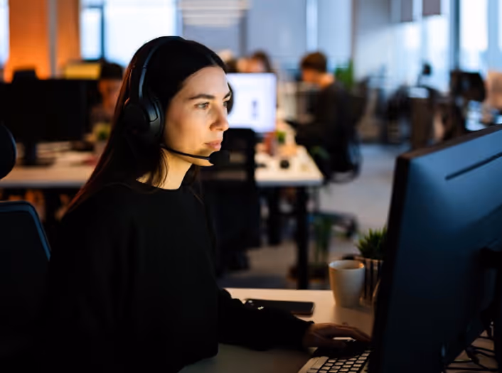 Woman wearing headset working intently on a computer in a modern office environment with colleagues in the background.