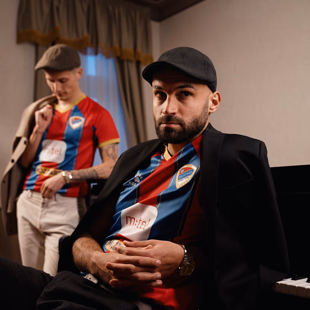Dva muškarca u crveno-plavim dresovima fudbalskog tima Borac, jedan sedi i nosi crnu kapu i sakoi preko dresa, drugi stoji u pozadini sa kapom i bež pantalonama.