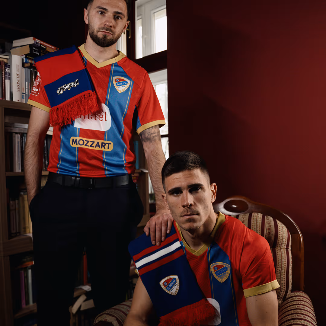 Dva mladića obučena u crveno-plave fudbalske dresove FK Borac sa šalovima u istoj boji, jedan stoji pored police s knjigama a drugi sedi na fotelji u prostoriji sa tamno crvenim zidom.