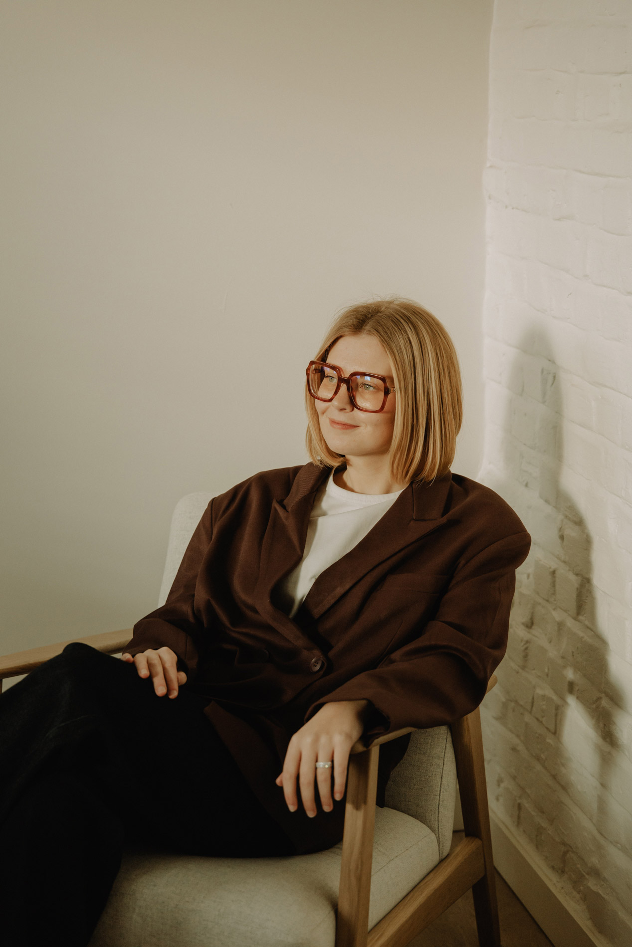 Vrouw met kort blond haar en grote bril zit ontspannen in een beige stoel tegen een witte bakstenen muur.